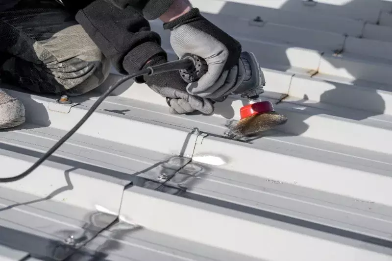 Työntekijä käyttää kulmahiomakonetta metallisen saumakattopinnan työstämiseen, kädet suojattu käsineillä ja työalueella näkyy kiinnikkeitä.