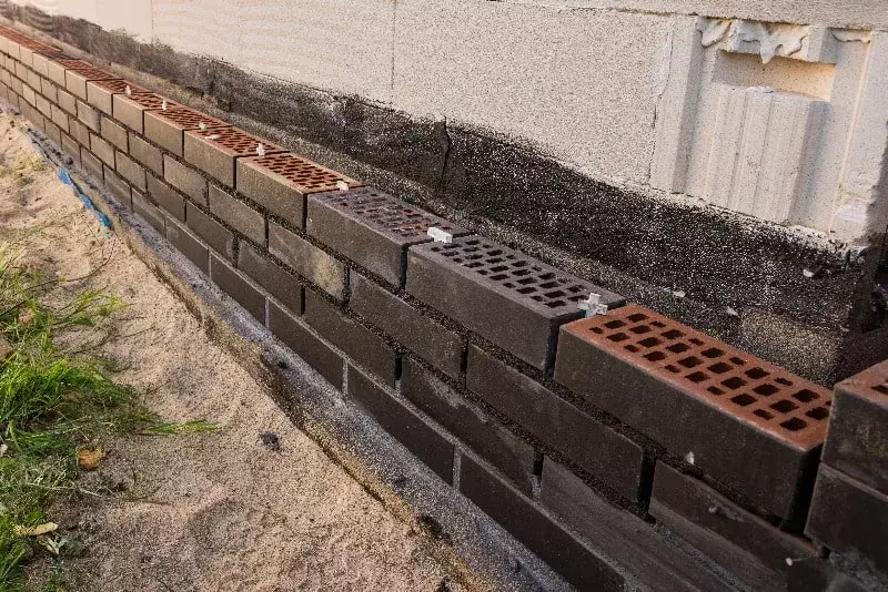 Keskeneräinen julkisivu tiiliestä jossa näkyy harmaita ja punaisia tiiliä ladottuna riveihin, taustalla betoniseinä.
