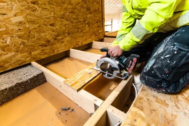 Rakennustyöntekijä sahaa lastulevyä käsipyörösahalla keltaisessa huomioväriasussa, ympärillä rakennusmateriaaleja.